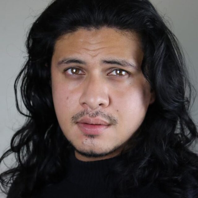 Close-up portrait of a man with long dark wavy hair and a moustache, looking directly at the camera