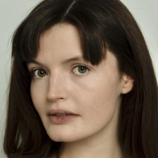 Close-up portrait of a woman with long dark hair, fringe, and green eyes, looking directly at the camera