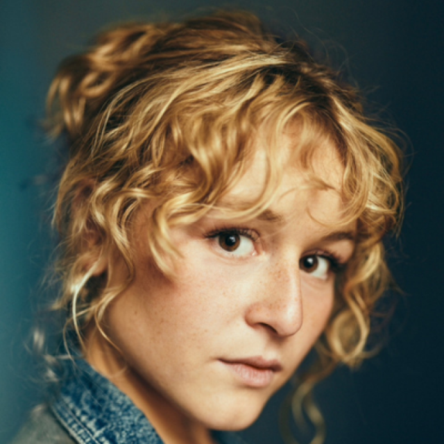Close-up portrait of a young woman with curly blonde hair, freckles and a denim jacket, gazing directly at the camera