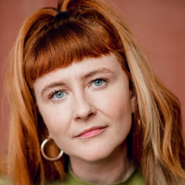 Close-up portrait of a woman with red hair and a fringe, wearing a hoop earring, looking directly at the camera