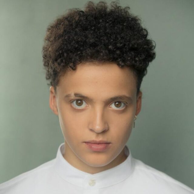 Portrait of a young person with curly hair, wearing a white shirt, looking directly at the camera