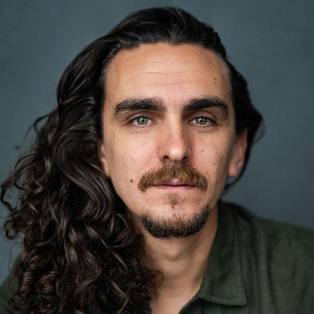 Portrait of a man with long curly dark hair and a beard, wearing a green shirt, against a grey background