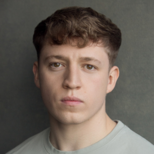 Headshot of a young man with curly brown hair, wearing a grey top, against a dark grey background
