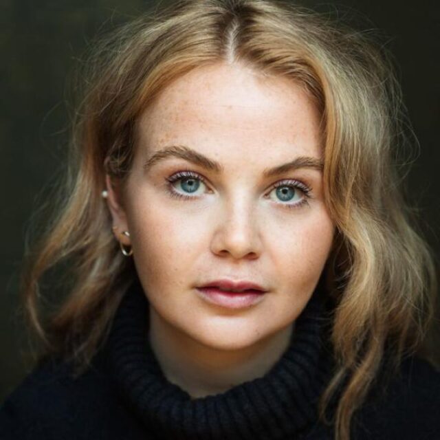 Close-up portrait of a young woman with wavy blonde hair, blue eyes and freckles, wearing a dark polo-neck jumper