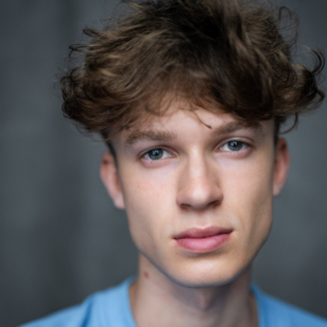 Close-up portrait of a young man with curly brown hair, wearing a light blue T-shirt, looking directly at the camera