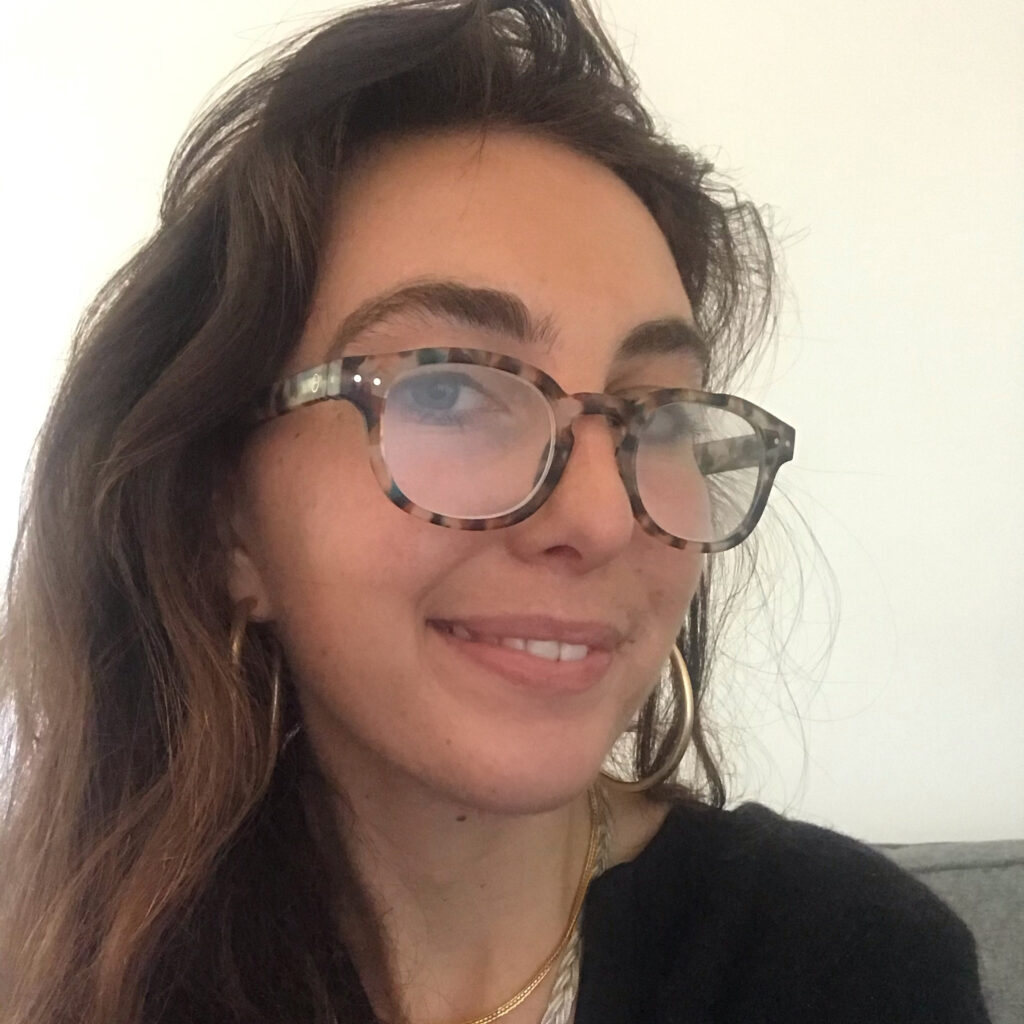 Close-up selfie of a smiling woman with long brown hair, wearing tortoiseshell glasses, hoop earrings and a black top