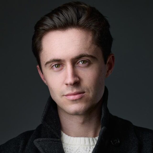 Studio headshot of a young man with brown hair and green eyes, wearing a dark coat over a cream jumper