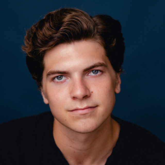 Headshot of a young man with wavy brown hair, wearing a black top against a dark blue background