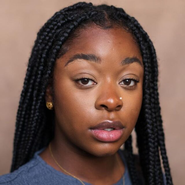 Close-up portrait of a young woman with box braids, gold earrings and necklace, looking directly at the camera