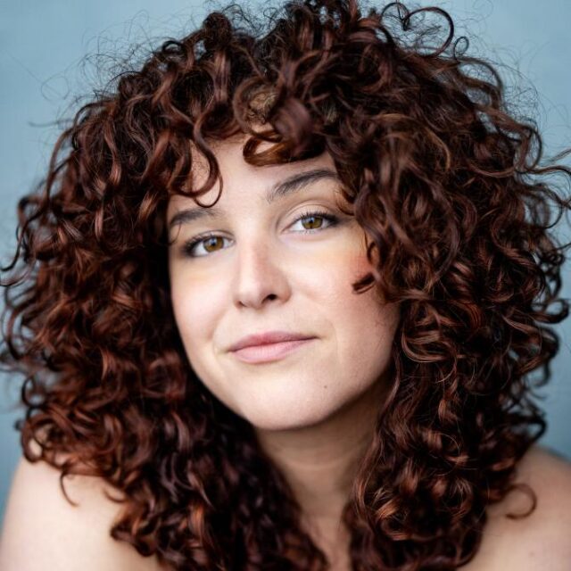 Woman with voluminous curly auburn hair, looking directly at the camera with a soft smile