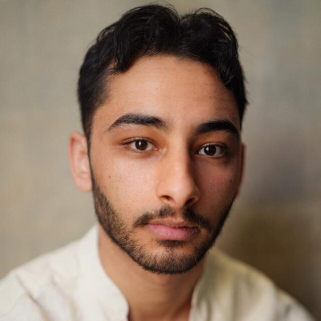 Close-up portrait of a young man with dark curly hair and a beard, looking directly at the camera