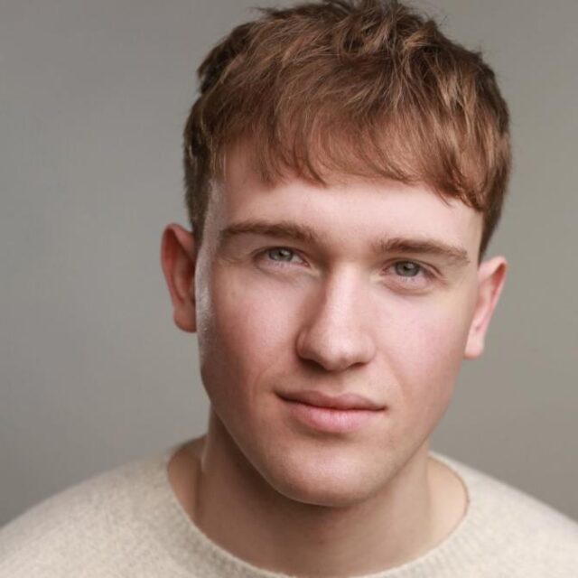 Headshot of a young man with reddish-brown hair, wearing a cream jumper, against a grey background