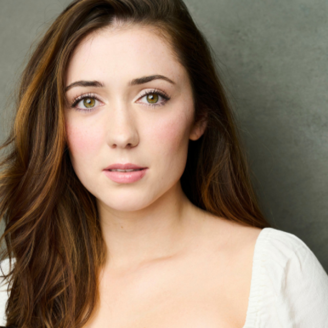 Close-up portrait of a young woman with long brown hair, wearing a white top, against a grey background