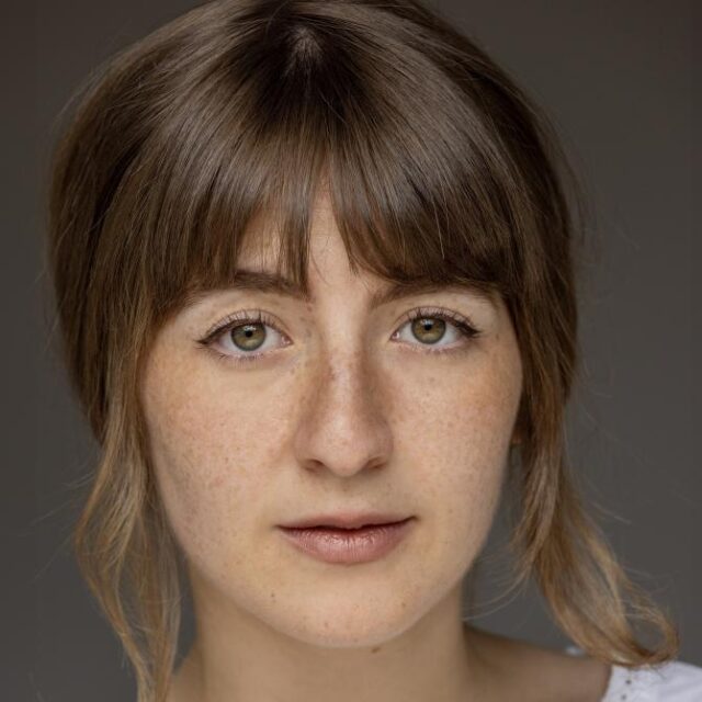 Close-up portrait of a young woman with brown hair and a fringe, green eyes and freckles, looking directly at the camera