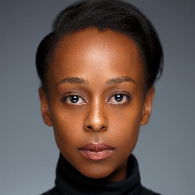 Close-up portrait of a woman with dark hair pulled back, wearing a black polo neck, looking directly at the camera