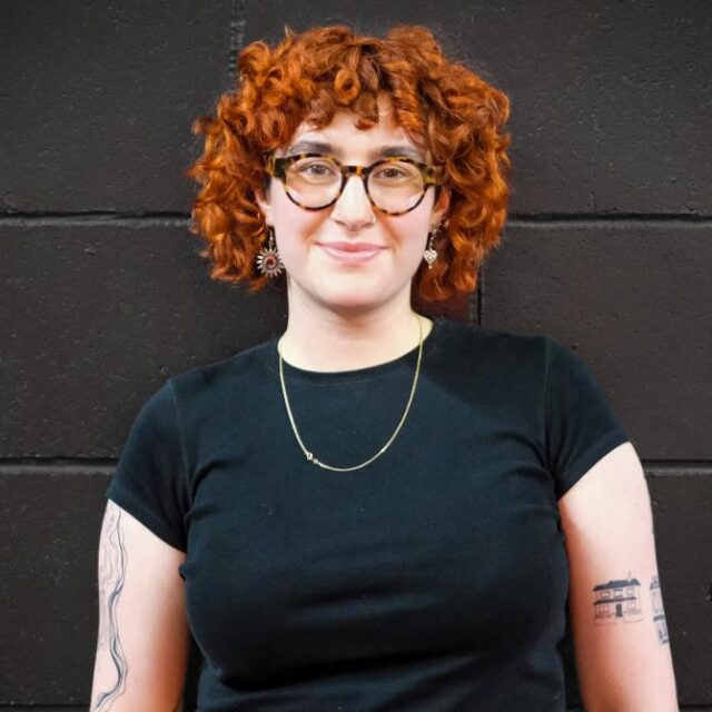Woman with curly red hair and tortoiseshell glasses, wearing a black t-shirt, smiling against a dark brick wall