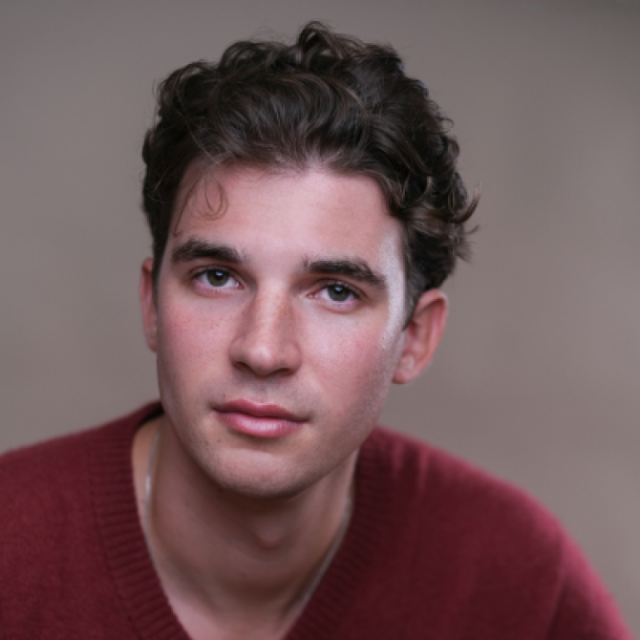 Headshot of a young man with curly dark hair, wearing a maroon jumper, looking directly at the camera
