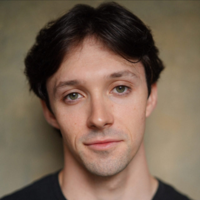 Headshot of a young man with dark, wavy hair and green eyes, wearing a dark T-shirt