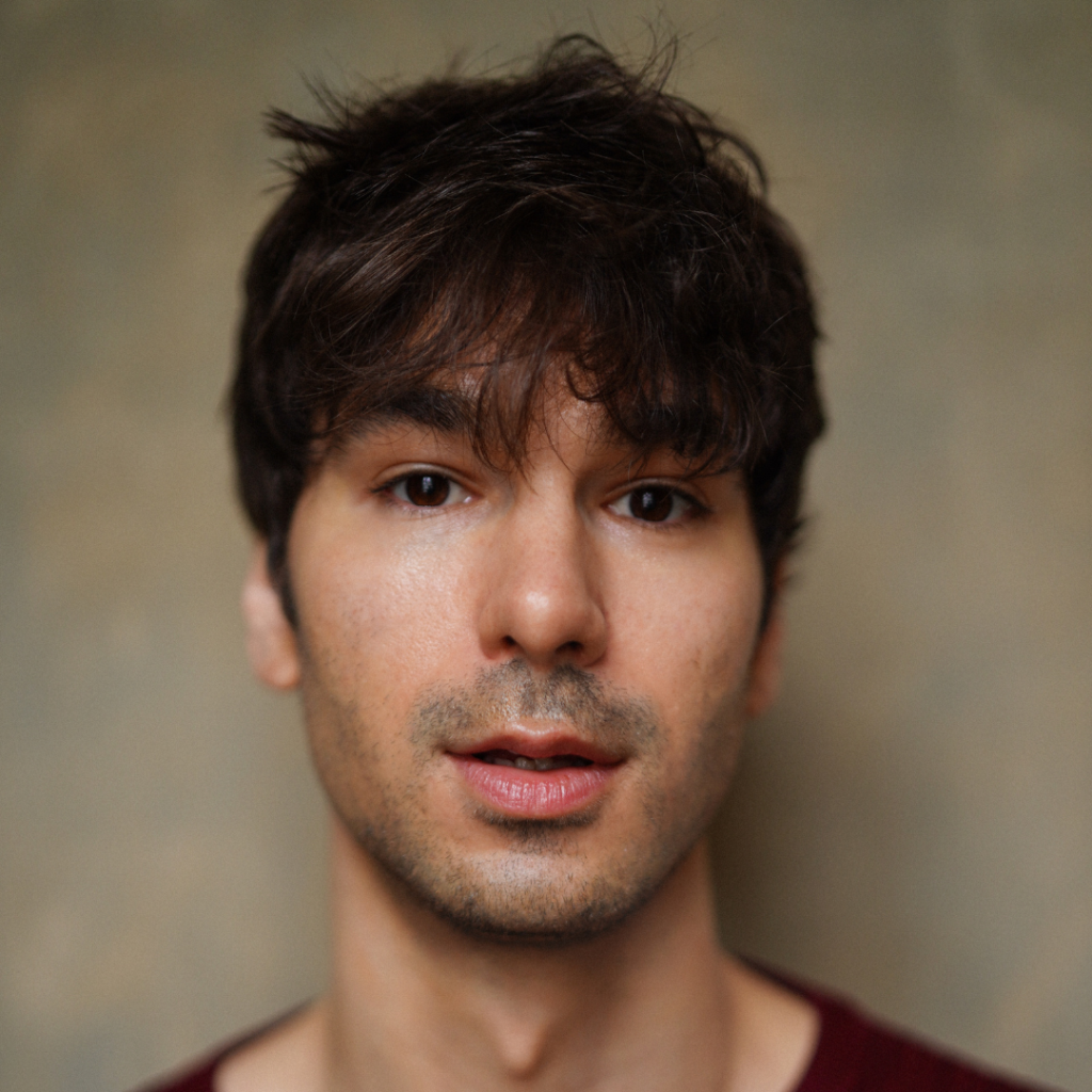 Close-up portrait of a young man with tousled dark hair and light stubble, looking directly at the camera