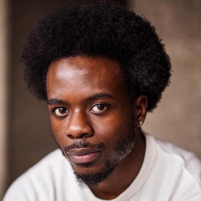 Close-up portrait of a man with an afro and beard, wearing a light-coloured top, looking directly at the camera
