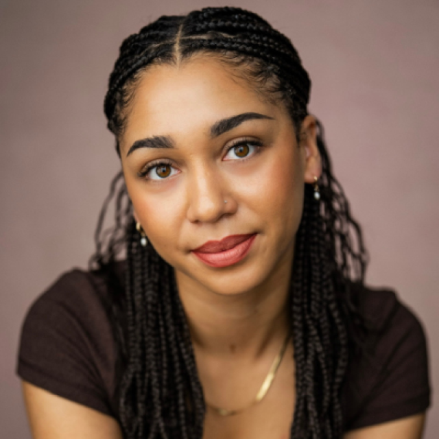 Woman with braided hair wearing a gold necklace, looking directly at the camera