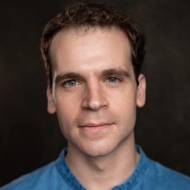 Portrait of a man with curly brown hair and blue eyes, wearing a denim shirt, against a dark background