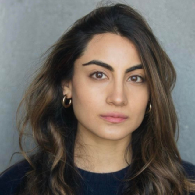 Headshot of a woman with long wavy brown hair, wearing gold hoop earrings and a dark jumper