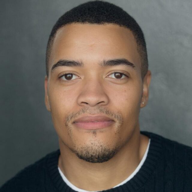 Headshot of a man with short hair and light stubble, wearing a dark jumper, against a grey background
