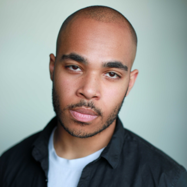 Bald man with a short beard, wearing a dark jacket over a white top, looking directly at the camera