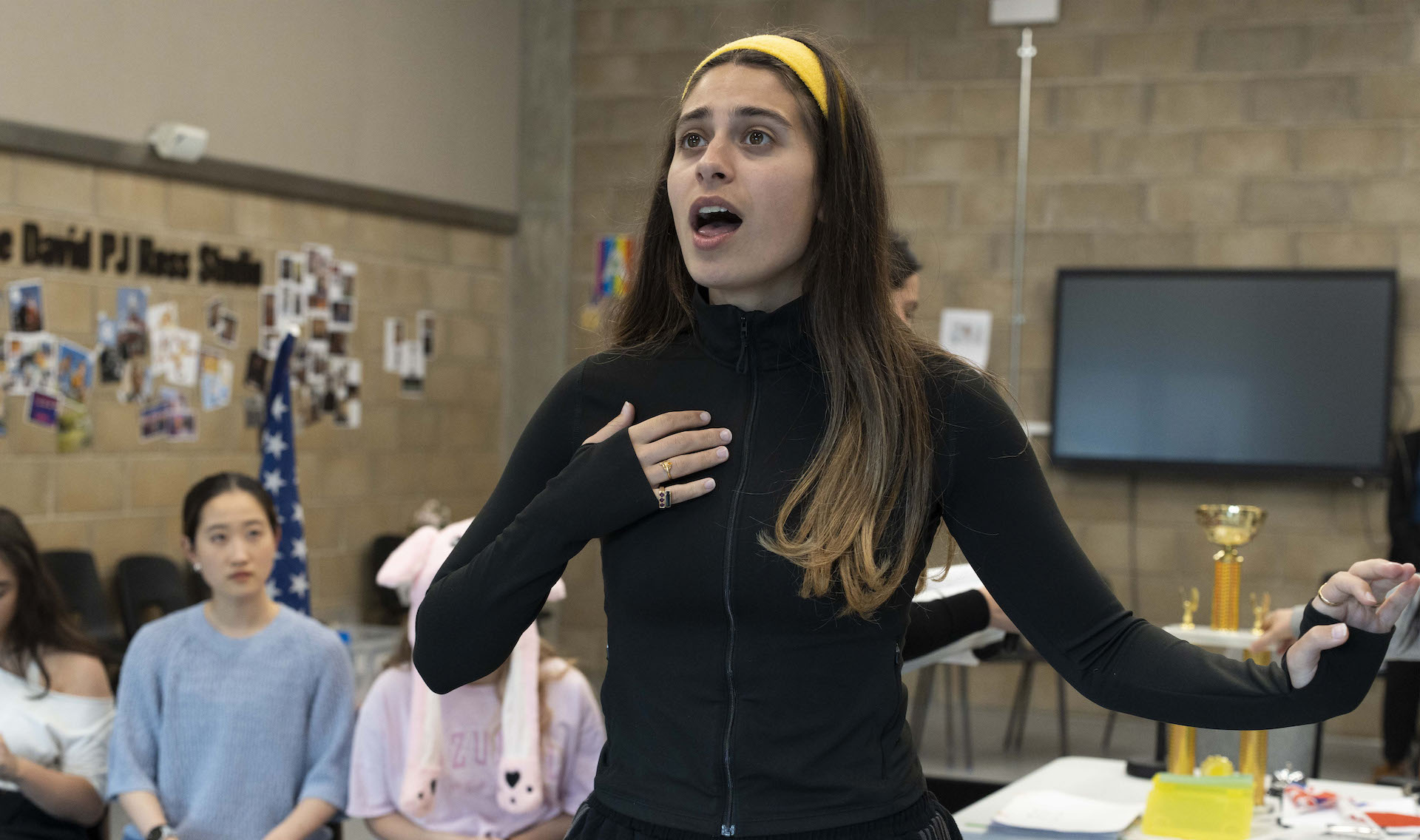Student performs a monologue with hand on chest during a drama class rehearsal, watched by seated classmates