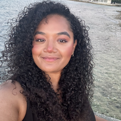 Woman with curly dark hair smiling at the camera, with the sea in the background