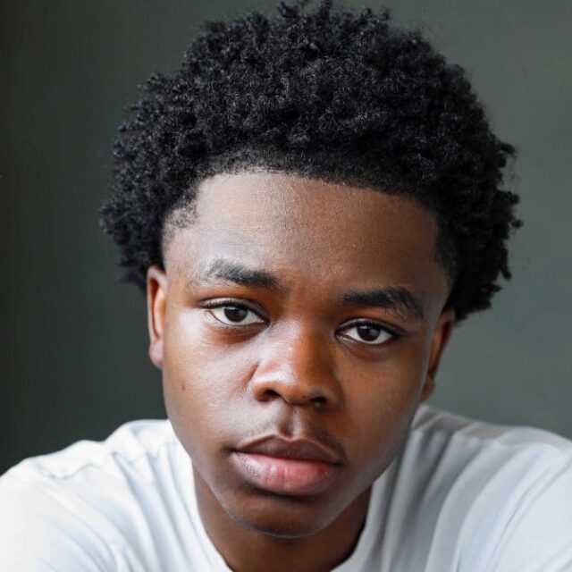 Close-up portrait of a young man with short curly hair, wearing a white top, looking directly at the camera