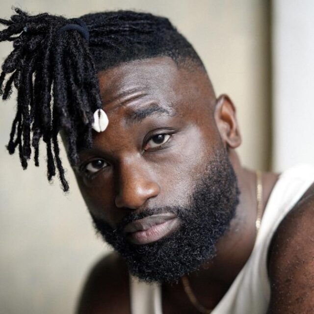 Man with locs tied up and a shell bead, beard, wearing a white vest and gold chain, looking at camera
