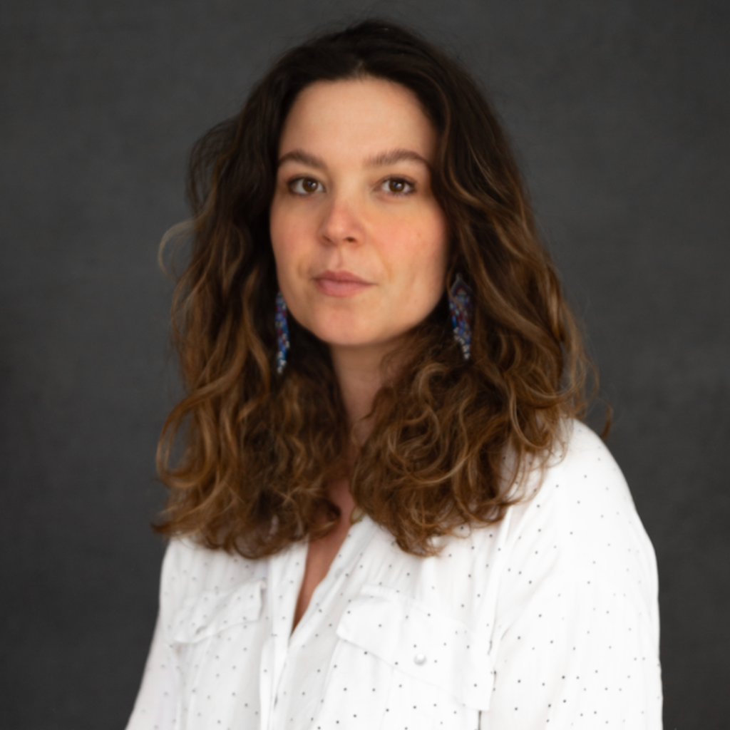 Portrait of a woman with long curly brown hair, wearing beaded earrings and a white spotted blouse