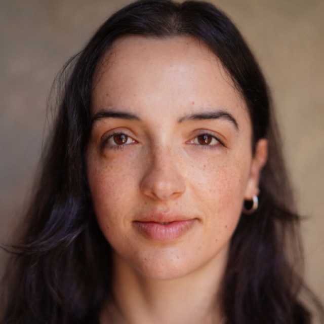 Close-up portrait of a woman with dark hair, freckles and a small hoop earring, looking directly at the camera
