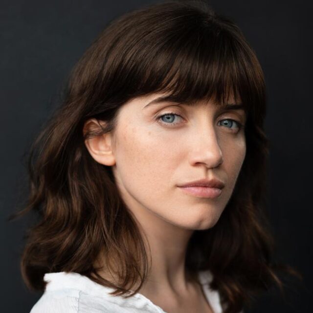 Close-up portrait of a woman with wavy brown hair and fringe, looking directly at the camera against a dark background