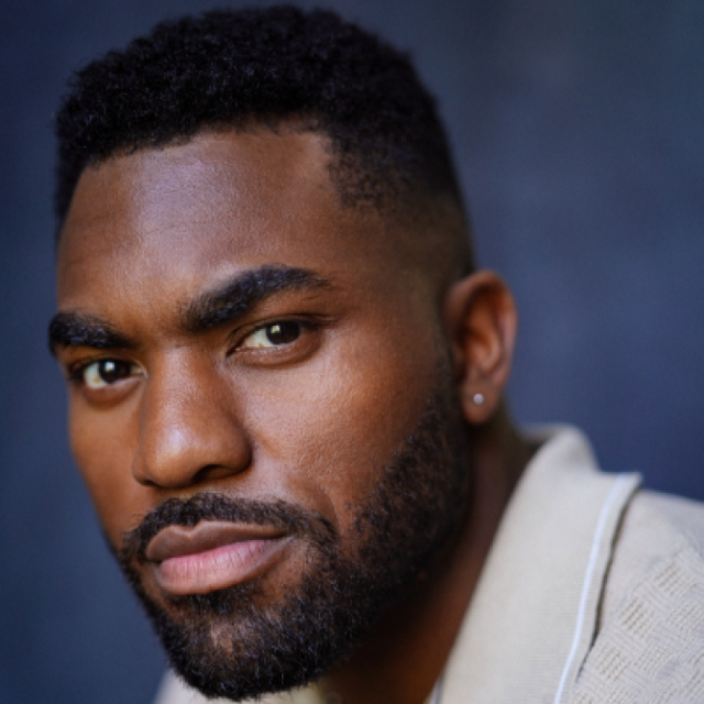 Close-up portrait of a man with short hair and a beard, wearing a cream-coloured jacket, against a dark background