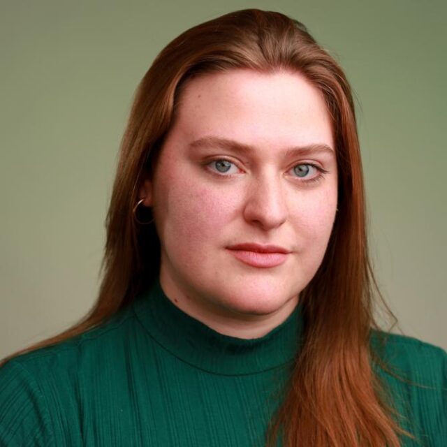 Portrait of a woman with long red hair wearing a green polo-neck top, against a green background