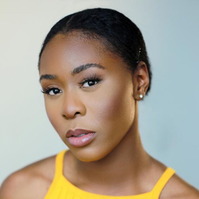 Close-up portrait of a woman with slicked-back hair, wearing a yellow top, gazing at the camera