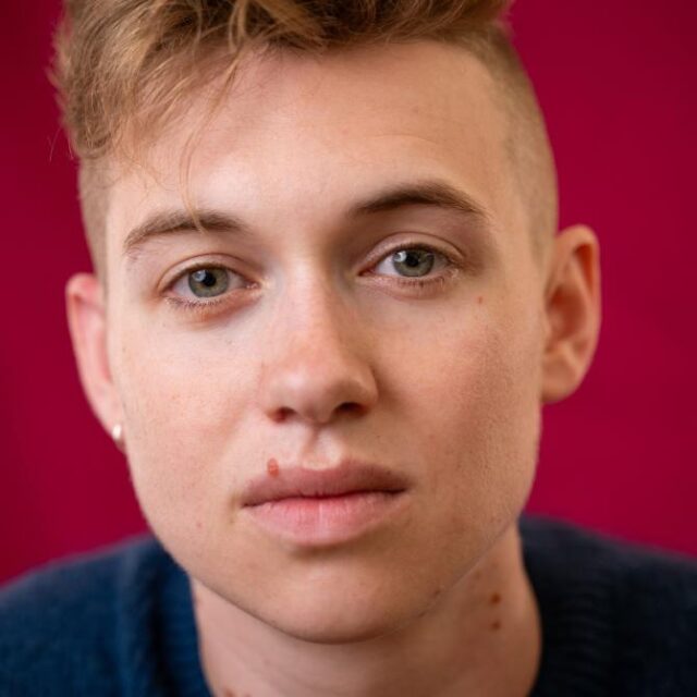 Close-up portrait of a young person with short, tousled reddish-blonde hair against a deep red background