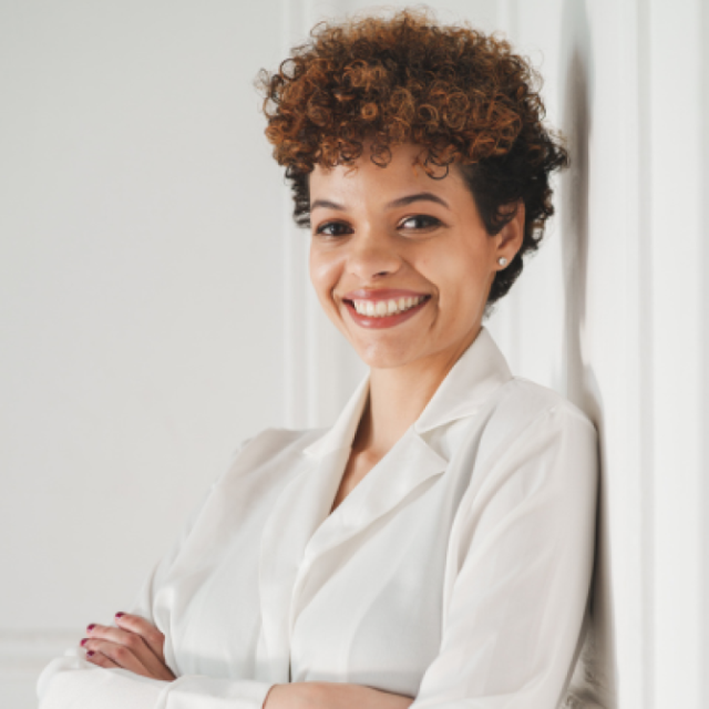 Smiling woman with curly hair, wearing a white blazer, standing with arms crossed