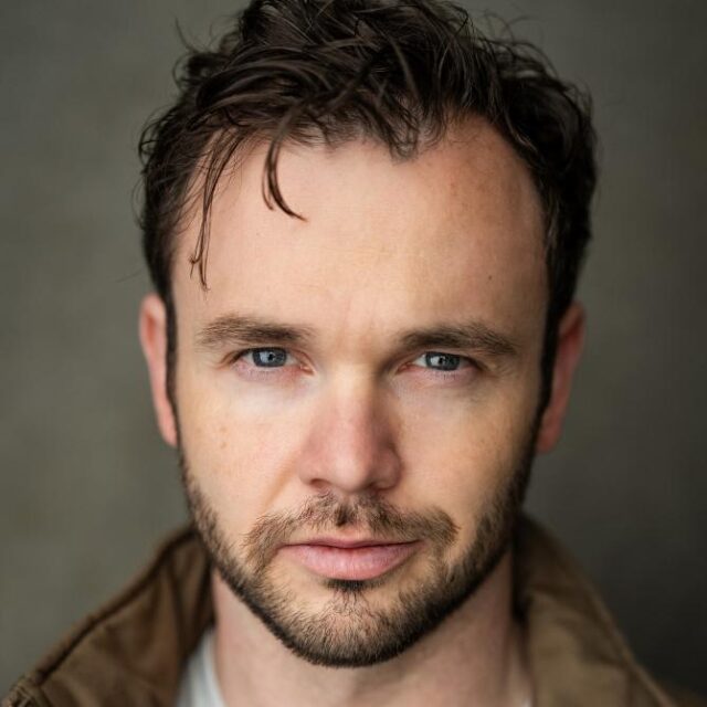 Close-up headshot of a man with wavy brown hair, blue eyes and a short beard, wearing a brown jacket