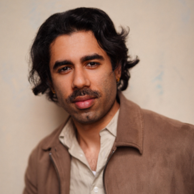 Portrait of a man with dark wavy hair and a moustache, wearing a tan jacket over a beige shirt