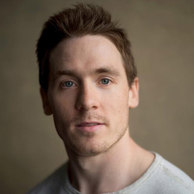 Portrait of a young man with short brown hair and blue eyes, wearing a light grey top