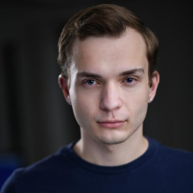 Close-up portrait of a young man with brown hair and earrings, wearing a navy top, looking directly at the camera