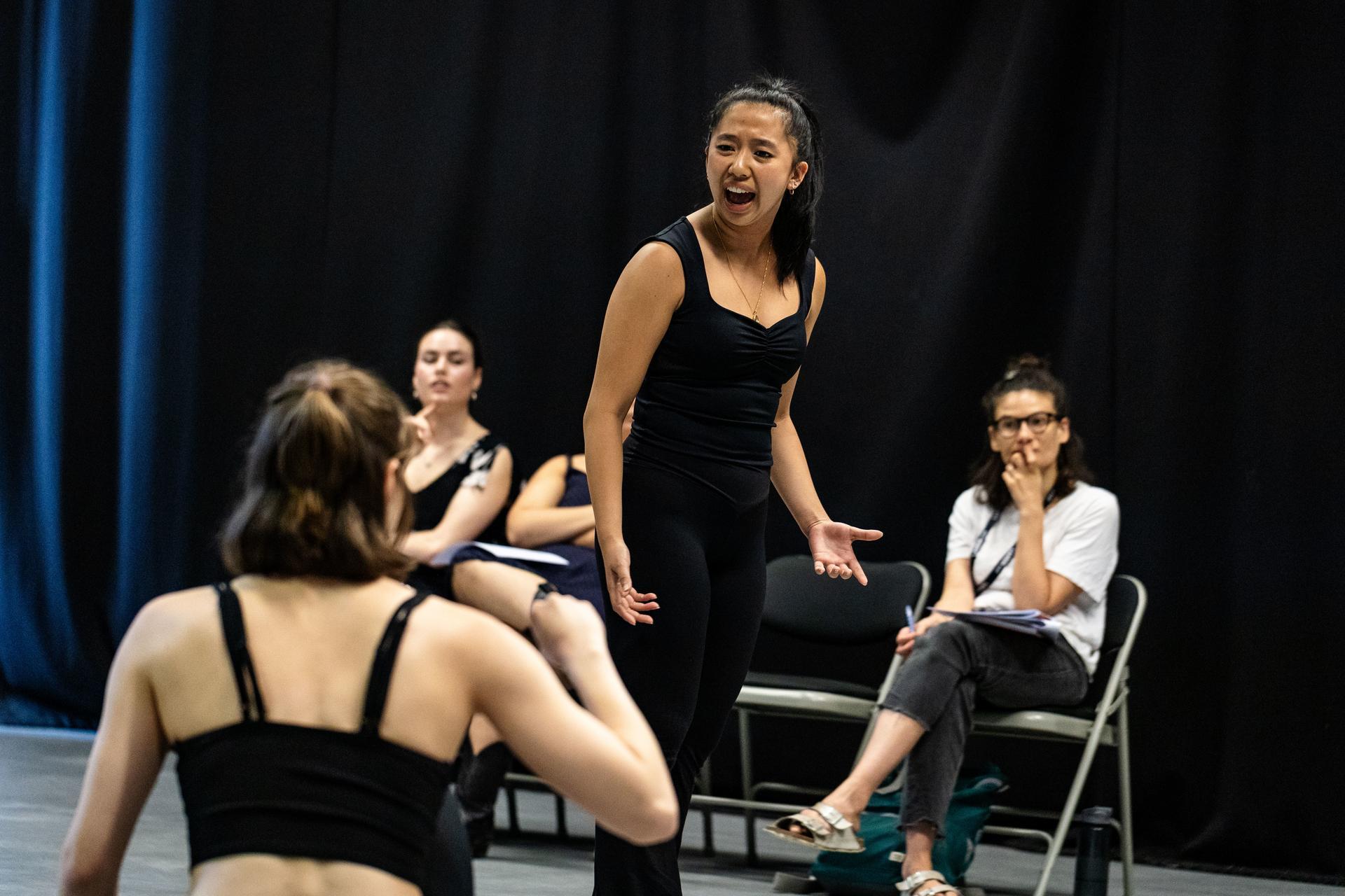 A performer in black rehearses expressively with an open gesture, while others watch and an observer takes notes nearby