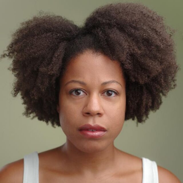 Close-up portrait of a woman with voluminous curly afro hair styled in two puffs, looking directly at the camera with a neutral expression