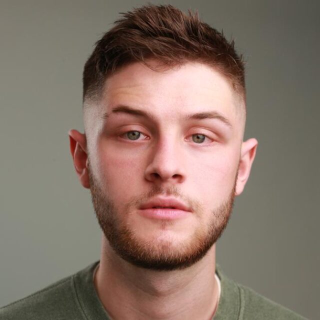 Close-up portrait of a young man with short brown hair and a beard, wearing a green top
