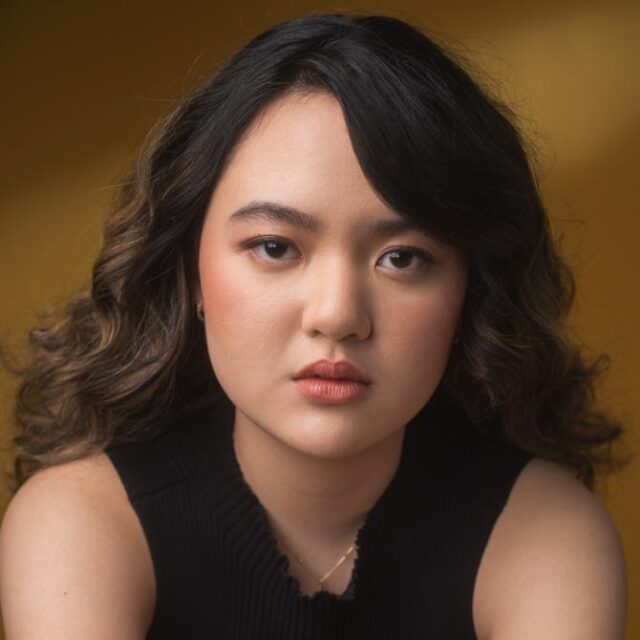 Close-up portrait of a young woman with curly dark hair, wearing a black top and looking directly at the camera