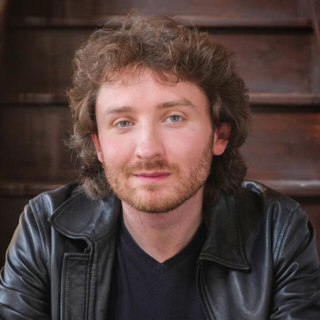 Man with curly brown hair and beard, wearing a leather jacket, seated against a wooden staircase backdrop